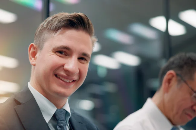 Ein junger Angestellter im Anzug lächelt im Büro in die Kamera